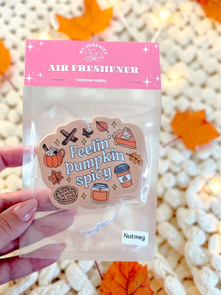 A hand holds a clear plastic bag containing a novelty air freshener shaped like a pumpkin slice. The top of the packaging has a pink header reading 'AIR FRESHIENER' with the brand name 'My Serenity' visible.
