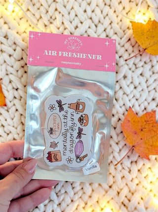 A hand holds a silver foil packet of 'Air Freshener' by the brand Serenity, featuring a pink header card and a vanilla-flavored sticker sheet with cute illustrations, set against a cozy white knit blanket with warm fairy lights and autumn leaves.