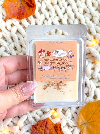A hand holds a clear plastic clamshell package of wax melts over a chunky white knit blanket with scattered autumn leaves and warm string lights. The label reads 'mentally at the dragonfly inn' and 'Pumpkin Cinnamon Rolls'.