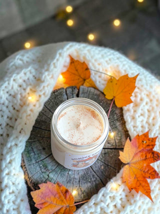 A jar candle labeled 'Cinnamon Spice' sits on a rustic wood slice, surrounded by a chunky white knit blanket and orange autumn maple leaves with warm fairy lights in the background.