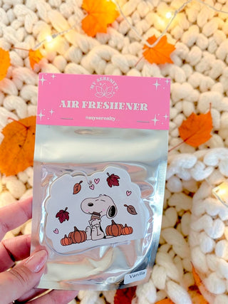 A hand holds a packaged Snoopy air freshener in a clear plastic bag with a pink header card. The air freshener features Snoopy sitting on pumpkins with autumn leaves and hearts.
