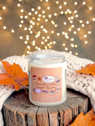 A glass jar candle labeled 'Mentally at the Dragonfly Inn' sits on a rustic wooden stump, wrapped in a white knit blanket with orange autumn leaves around it, against a warm background of glowing fairy lights.