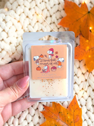 A hand holds a clear plastic clamshell package containing a wax melt or scented melt bar with a peach label featuring Snoopy illustrations and the phrase 'I love fall most of all.' The background shows a cream knit blanket and orange autumn leaves.