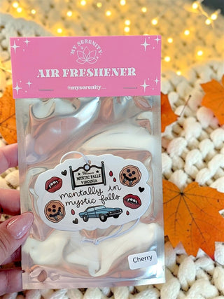 A hand holds a clear plastic bag of air freshener sachets with a pink header card reading 'AIR FRESHENER' and the brand name 'The Serenity'. The sachets inside are pale pink and white, and the front has a decorative sticker with illustrations and the text 'mentally in a mystic valley'.