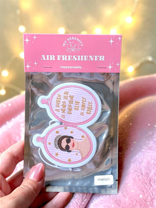 A hand holding a packaged air freshener featuring two pink and white oval stickers with text and an illustration, set against a warm, blurred background with bokeh lights and a pink blanket.