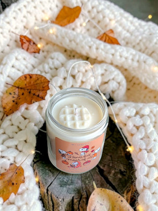 A small glass jar candle with white wax sits on a rustic wood slice, surrounded by a chunky cream knit blanket, scattered autumn leaves, and warm fairy lights. The candle label is peach-colored with cute cartoon illustrations and text that appears to read 'Pumpkin Spice' and 'Warm & Sweet.