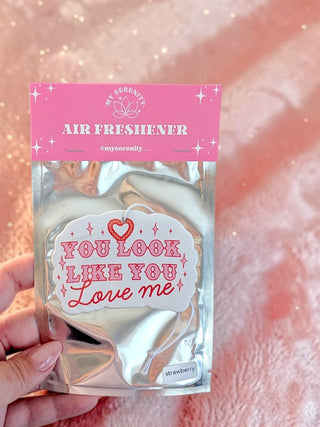 A hand holds a silver foil pouch of air freshener with a pink header card that reads 'MY SERENITY AIR FRESHENER'. The front label says 'YOU LOOK LIKE YOU Love me' with a small red heart.