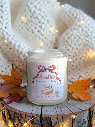 A glass jar candle labeled 'Sookie's Cinnamon Rolls' sits on a rustic wood slice with autumn leaves and warm fairy lights, in front of a cream knit blanket. Gilmore inspired