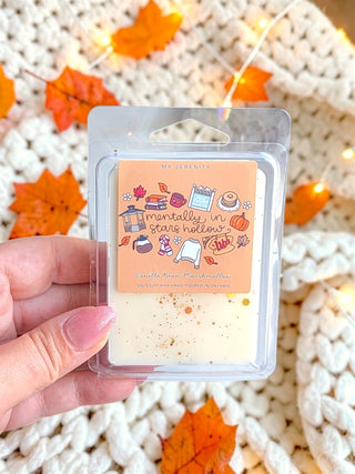 A hand holds a clear plastic clamshell package containing a small wax melt or scented melt bar with a peach label that reads 'mentally in stars holes' and 'Candle Bean Marshmallow.' The background is a cozy white knit blanket with scattered orange autumn leaves and warm string lights.