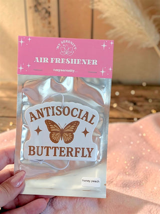 A hand holds a silver resealable pouch containing an air freshener. The top of the pouch is pink with white text reading 'MY SERENITY AIR FRESHENER', and the front label is white with brown text reading 'ANTISOCIAL BUTTERFLY' and a butterfly illustration.