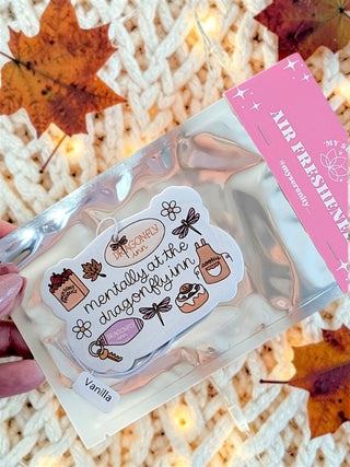 A hand holds a clear plastic pouch containing a vanilla air freshener sticker set, with a pink header card reading 'AIR FRESHENER' and 'Vanilla'. The background shows a cozy white knit blanket with scattered autumn leaves and warm string lights.
