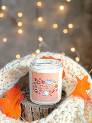 A white candle in a clear glass jar sits on a small wood slice, wrapped in a cream knit blanket with orange autumn leaves. The candle label reads 'Mia's Treasures' and 'Essentially in static hollers' with fall-themed illustrations.
