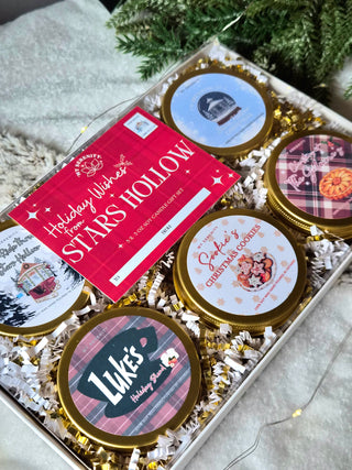A cozy flat lay of the “Holiday Wishes from Stars Hollow” candle gift set from My Serenity Shop. Five 3oz gold-lidded tins are arranged with twinkle lights, greenery, and festive accents. Each label features Gilmore Girls–inspired names like Christmas at the Gilmores, Luke’s Holiday Blend, Sookie’s Christmas Cookies, Sleigh Ride Through Stars Hollow, and I’m Dreaming of a Stars Hollow Christmas. Warm, nostalgic tones of gold, cream, and burgundy evoke small-town holiday charm.