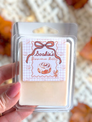 A hand holding a clear plastic clamshell package of My Serenity Soakie's Cinnamon Rolls wax melts, showing the pale cream wax inside and the decorative pink and white label.