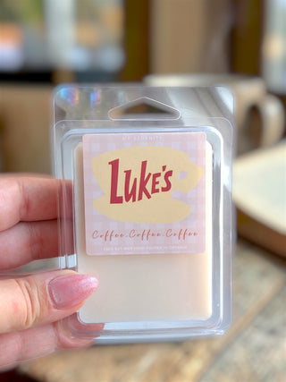 A hand holding a packaged wax melt in a clear plastic clamshell. The label reads 'Luke's Coffee - Coffee - Coffee' with a cream-colored wax melt visible inside.
