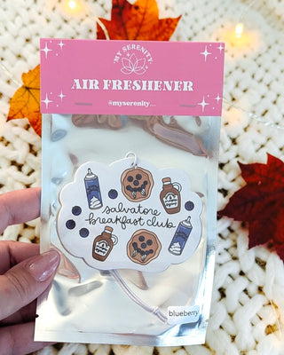 A hand holds a packaged air freshener shaped like a cloud, with a pink header card reading 'MY SERENITY AIR FRESHENER' and the scent name 'salvatore breakfast club' on the front. The package is in a shiny silver pouch over a white knit blanket, with autumn leaves and warm string lights in the background.