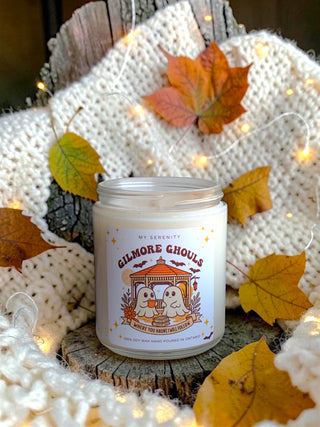 A white candle in a clear glass jar sits on a rustic wood slice, surrounded by a chunky cream knit blanket, autumn leaves, and warm fairy lights. The label reads 'MY SERENITY' and 'GILMORE GHOULS'.