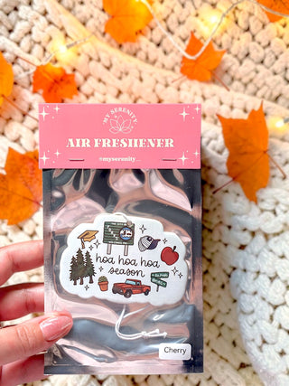 A hand holds a packaged cherry scented air freshener by My Serenity. The product is in a clear plastic bag with a pink header card and a white cloud-shaped sticker featuring a cherry tree and 'hoa hoa hoa season' text.
