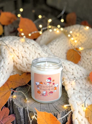 A small glass jar candle with a peach label featuring cartoon characters and the phrase 'I love fall means to all,' styled on a wooden stump with autumn leaves, a chunky cream knit blanket, and warm fairy lights in the background.